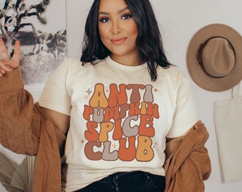 May include: A woman wearing a white t-shirt with the text "ANTI PUMPKIN SPICE CLUB" in a retro font. The text is in shades of orange, brown, and gray. She is wearing a brown jacket and has long dark hair.