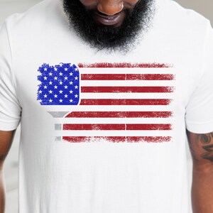 May include: White t-shirt with a distressed American flag design featuring a pickleball paddle in the upper left corner.
