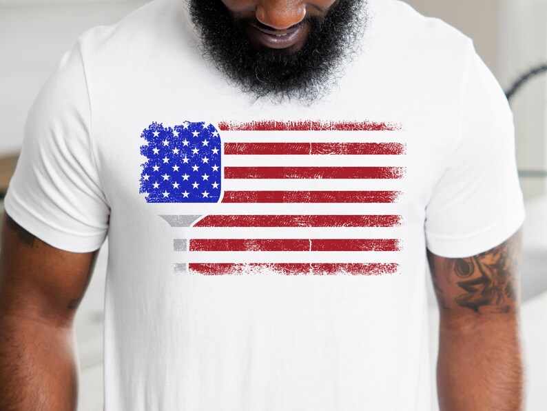 May include: White t-shirt with a distressed American flag design featuring a pickleball paddle in the upper left corner.