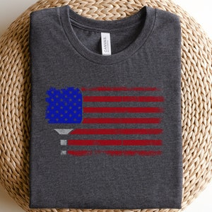 May include: A dark heather grey t-shirt with a distressed American flag design featuring a pickleball paddle in the upper left corner.