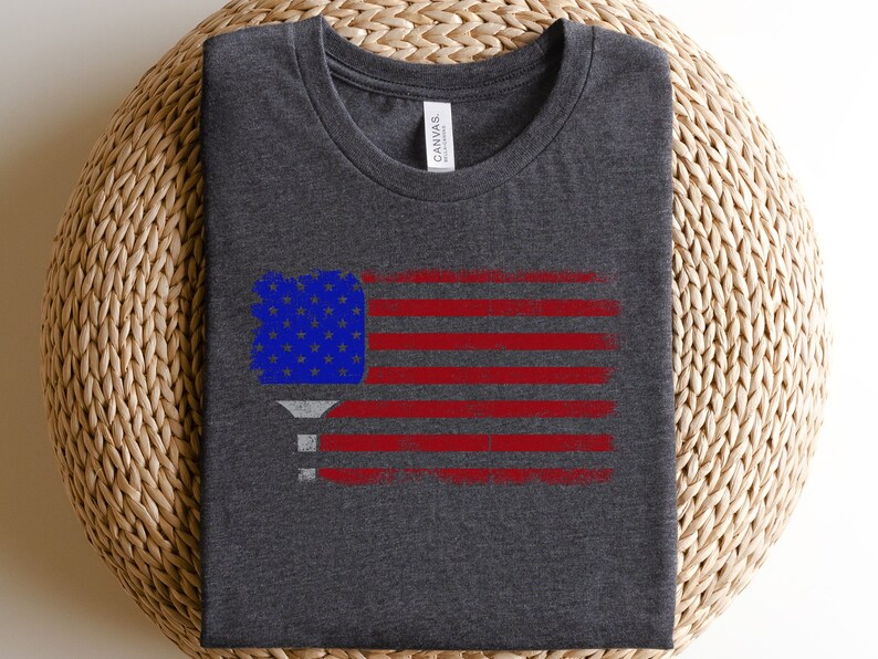 May include: A dark heather grey t-shirt with a distressed American flag design featuring a pickleball paddle in the upper left corner.