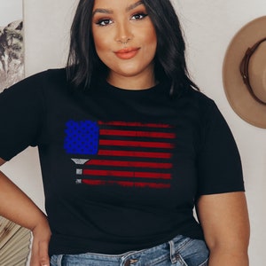 May include: A black t-shirt with a distressed American flag design featuring a blue and white pickleball paddle. The flag is red, white, and blue.
