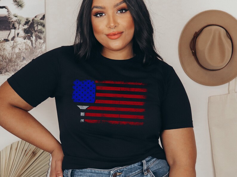 May include: A black t-shirt with a distressed American flag design featuring a blue and white pickleball paddle. The flag is red, white, and blue.