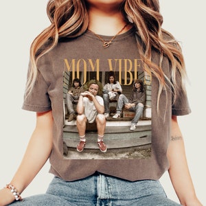 May include: A brown t-shirt with a graphic print of four people sitting on a porch. The text "MOM VIBES" is printed in gold above the image.