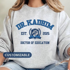 May include: Gray sweatshirt with blue text that reads "Dr. Kadhim Est. 2025 Doctor of Education" with a blue lion crest. The text "Customizable" is at the bottom of the sweatshirt.