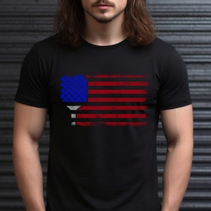 May include: A black t-shirt with a distressed American flag design featuring a pickleball paddle in the center.