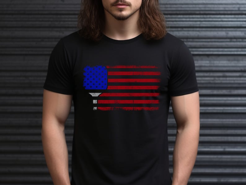 May include: A black t-shirt with a distressed American flag design featuring a pickleball paddle in the center.