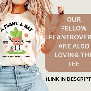 May include: A white t-shirt with a green plant cartoon wearing a brown pot and the text "A Plant A Day" with the words "Grow More" and "Worry Less" on either side of the plant. The text "Keeps the Anxiety Away" is below the plant. The text "Our Fellow Plantroverts Are Also Loving This Tee (Link in Description)" is on a brown background.