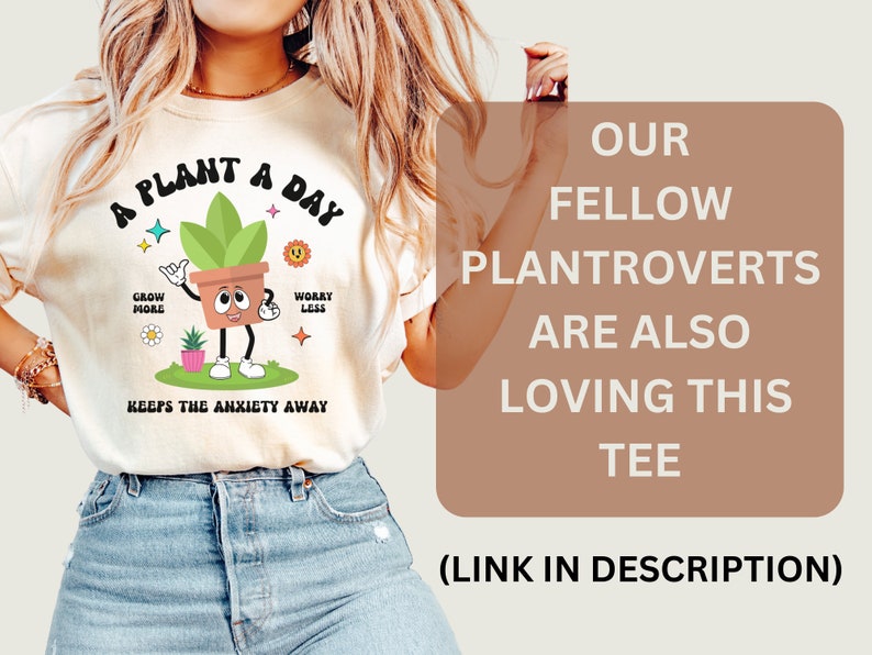 May include: A white t-shirt with a green plant cartoon wearing a brown pot and the text "A Plant A Day" with the words "Grow More" and "Worry Less" on either side of the plant. The text "Keeps the Anxiety Away" is below the plant. The text "Our Fellow Plantroverts Are Also Loving This Tee (Link in Description)" is on a brown background.
