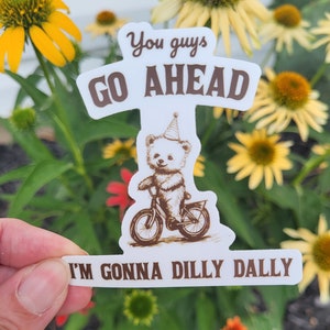 May include: White sticker with brown text that reads "You guys go ahead I'm gonna dilly dally". A cartoon bear wearing a party hat rides a bicycle.