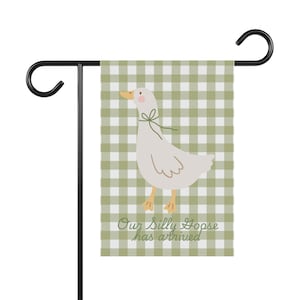 May include: A garden flag featuring a cartoon goose with a green ribbon around its neck, set against a green and white checkered background. The text "Our Silly Goose has arrived" is printed at the bottom. The flag is attached to a black metal stand.