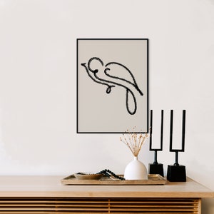 以下が含まれることがあります： 白い壁に黒い額縁に入った、枝にとまった鳥の白黒線画が飾られています。その絵は、木製のテーブルの上に置かれた、ドライフラワーの入った花瓶、黒い金属製の燭台2つ、そしてボウルとビーズが入ったトレイの上にあります。