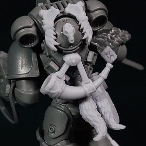 Puede incluir: Miniatura de plástico gris de una figura futurista blindada, posiblemente para juegos de mesa. La figura está posada sobre una base marrón texturizada, con detalles intrincados en la armadura y las armas. La figura sostiene un objeto peludo.