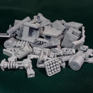 Puede incluir: Un montón de piezas de modelo de plástico gris claro sin montar. Los componentes incluyen bloques rectangulares, cilindros y otras formas geométricas, lo que sugiere un tema mecánico o industrial. Las piezas están esparcidas sobre una superficie verde oscuro.