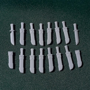 Puede incluir: Una colección de pequeñas espadas y fundas de juguete de color gris claro dispuestas sobre una superficie verde oscuro. Las espadas tienen una hoja curva y un mango rectangular. Las fundas son rectangulares con un pequeño mango.