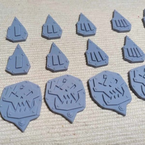 Peut inclure: Un ensemble de 12 pièces de jeu de guerre miniatures en plastique gris. Les pièces présentent différents motifs, notamment des crânes, des pointes et des formes géométriques.