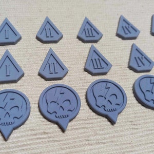 Puede incluir: Un conjunto de 15 piezas de juego de plástico gris. Las piezas tienen forma de triángulos y círculos. Los triángulos tienen los números romanos I, II y III. Los círculos tienen una calavera con un rayo.