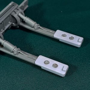 May include: A close-up of a grey mechanical component with two light grey rectangular pads, each with two silver circular details. The component is set against a dark green background.