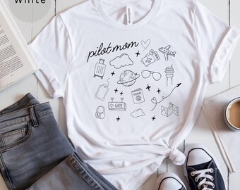 May include: White t-shirt with a black graphic design that says "pilot mom" and includes various travel-themed icons like a suitcase, passport, airplane, and clouds.