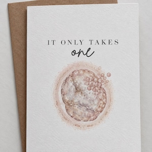 Può includere: Un biglietto d'auguri bianco con una busta marrone che riporta il testo IT ONLY TAKES one in inchiostro nero. Sotto il testo c'è un'illustrazione ad acquerello di una cellula uovo circondata da spermatozoi in tonalità rosa e beige.