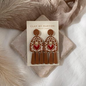 May include: A pair of brown clay earrings with a red heart design and gold accents. The earrings are dangle style and have a tassel-like design. The earrings are on a white card with the text "CLAY BY MARYORI handmade jewelry".