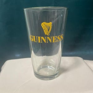 May include: A clear pint glass with the Guinness logo in yellow. The logo features a harp and the word "Guinness" in a bold, sans-serif font. The glass is empty and has a slightly tapered shape.
