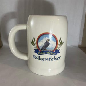 May include: Cream-colored ceramic beer stein with a large handle. The stein features a circular emblem with a bird, mountain, and wheat design, with the word "Falkenfelser" in blue script.