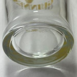 May include: Clear glass drinking glass with a thick, rounded base. The bottom of the glass has a slight yellow tint. The glass appears to be clean and in good condition. The image is a close-up of the bottom of the glass.
