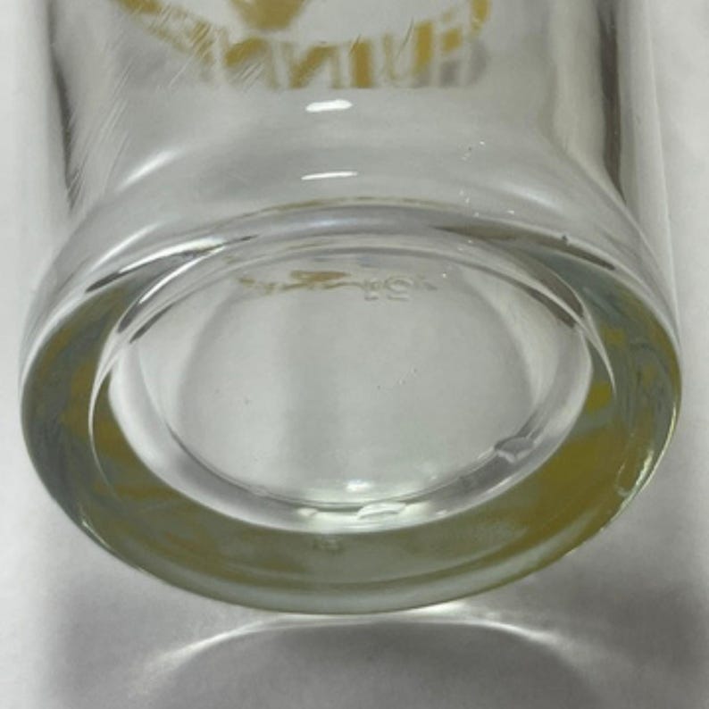 May include: Clear glass drinking glass with a thick, rounded base. The bottom of the glass has a slight yellow tint. The glass appears to be clean and in good condition. The image is a close-up of the bottom of the glass.