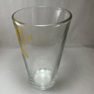 May include: A clear, tapered glass with a rounded rim. The glass has a yellow logo on the side. The glass is empty and sits on a white surface. The glass is designed for drinking.