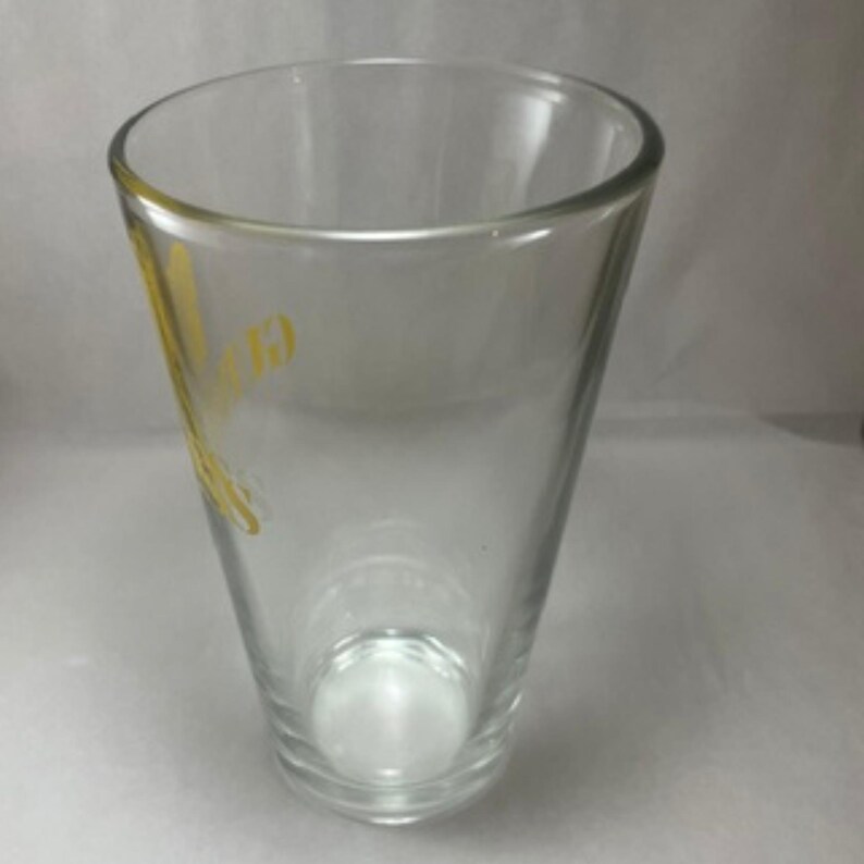 May include: A clear, tapered glass with a rounded rim. The glass has a yellow logo on the side. The glass is empty and sits on a white surface. The glass is designed for drinking.