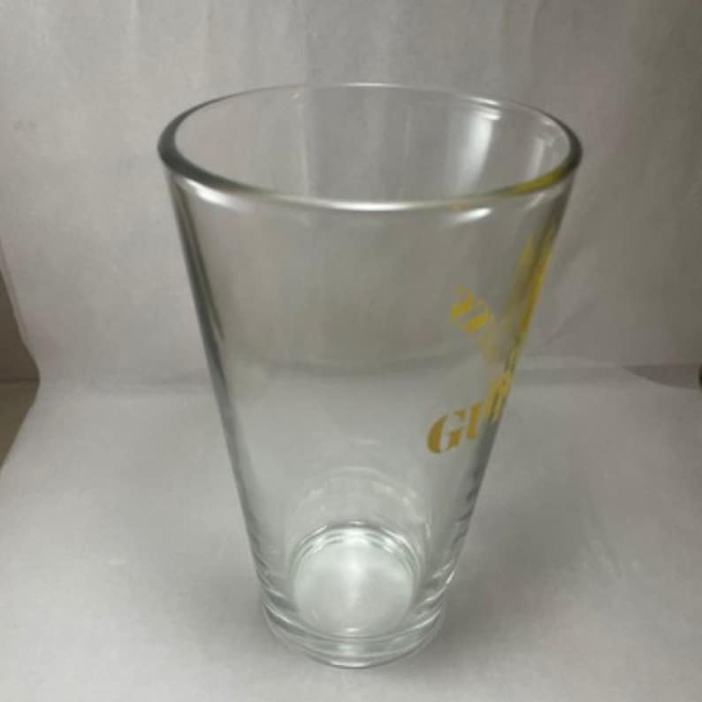 May include: A clear, tapered pint glass with a rounded rim. The glass has a yellow logo on the side. The glass is empty and sits on a white surface. The glass is designed for beverages.