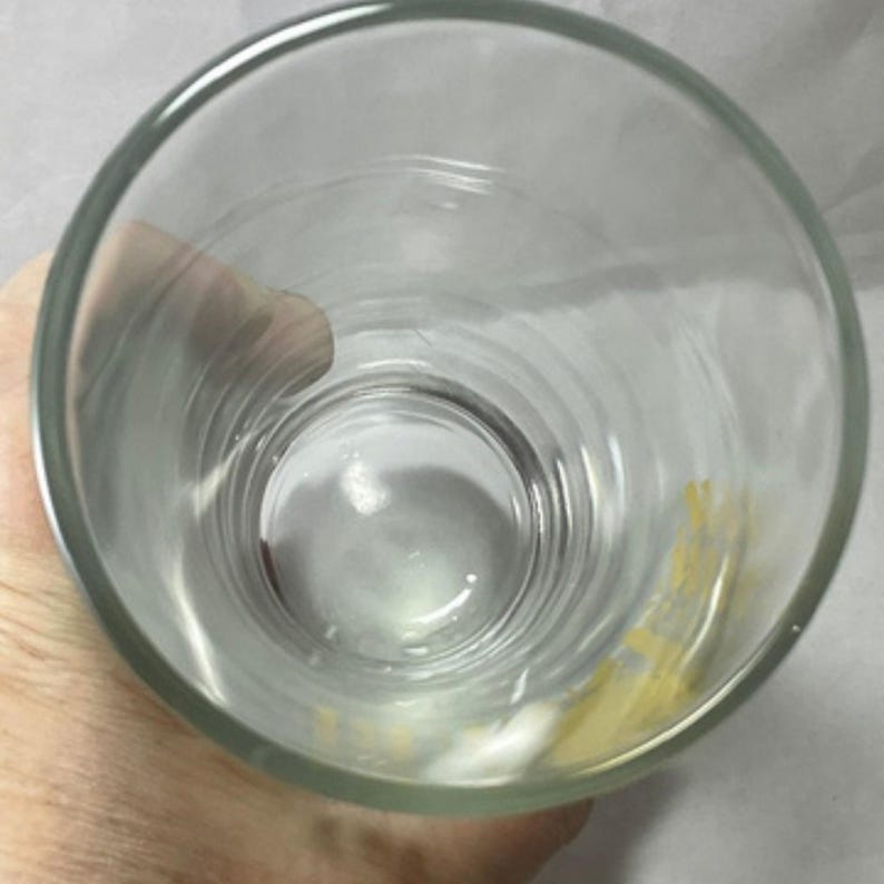 May include: A clear, cylindrical drinking glass with a thick rim. The glass is empty and appears clean, with slight discoloration on the interior. The image is taken from a top-down perspective, looking into the glass.