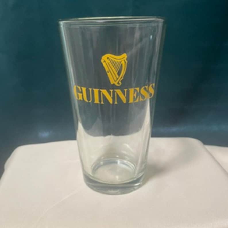 May include: A clear pint glass with the Guinness logo in yellow. The logo features a harp and the word "Guinness" in a bold, sans-serif font. The glass is empty and has a slightly tapered shape.