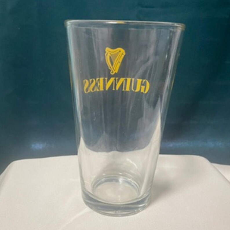 May include: A clear pint glass with the Guinness logo in yellow. The logo features a harp symbol and the Guinness name in a yellow, sans-serif font. The glass is empty and sits on a white surface against a dark teal background.