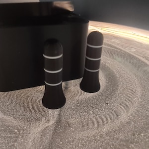 May include: A black sand art kit with a rectangular base and two cylindrical tools. The tools have white stripes. The sand is light-coloured and patterned with circular designs.