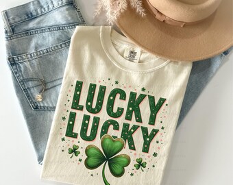 May include: A cream-colored t-shirt with the text "Lucky Lucky" in green and gold letters with a green shamrock design. The shirt is displayed on a denim background with a brown hat.