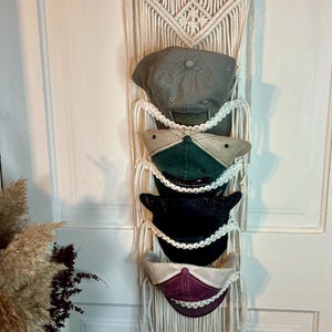 May include: A macrame hat organizer with five baseball caps. The caps are in various colors, including green, black, and gray. The organizer is made of cream-colored rope and hangs on a white door.