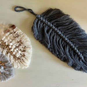 May include: A close-up of three macrame feather decorations. The largest is a dark gray, textured feather with a loop for hanging. Two smaller, fan-shaped pieces are off-white and gray, adorned with glitter and cinnamon sticks.