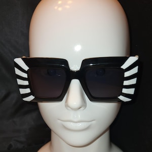 May include: Black square sunglasses with white striped accents on the frames. The sunglasses have dark lenses and are displayed on a white mannequin head against a black background. A stylish accessory.
