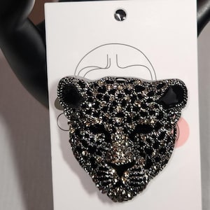 Può includere: Spilla a forma di testa di leopardo nera e argento. La spilla è realizzata con piccoli cristalli scintillanti e presenta accenti neri per orecchie e occhi. La spilla è attaccata a un cartoncino bianco con un'illustrazione nera.