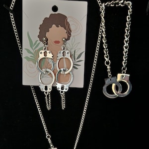 May include: A silver necklace and earrings set featuring a pair of handcuffs as the pendant and earring design. The necklace and earrings are on a white card with a drawing of a woman's face.