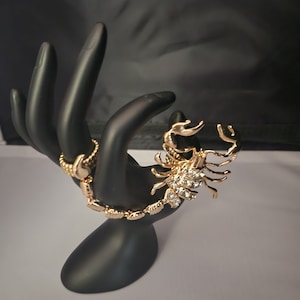 May include: A gold-coloured scorpion bracelet with clear crystal accents is displayed on a black hand-shaped jewellery holder. The scorpion's body is articulated, and the bracelet is set against a dark background.