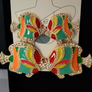 Puede incluir: Un par de pendientes de tono dorado con un diseño de pez colorido. Los peces están pintados en tonos de verde, amarillo, rojo y naranja. Los pendientes están adornados con brillantes transparentes.