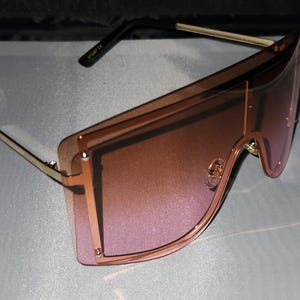 May include: Oversized sunglasses with a gold frame and pink gradient lenses. The frame has a geometric shape with a double layer design.