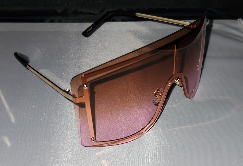 May include: Oversized sunglasses with a gold frame and pink gradient lenses. The frame has a geometric shape with a double layer design.