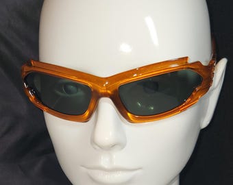 May include: Amber-colored sunglasses with a dark green lens. The sunglasses have a curved, wraparound design.