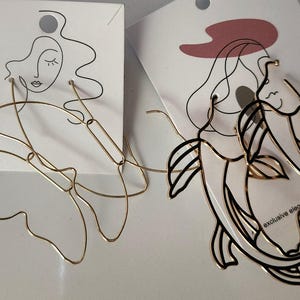 May include: Two pairs of gold earrings. One pair is a simple, abstract design. The other pair is a more detailed design of two fish.