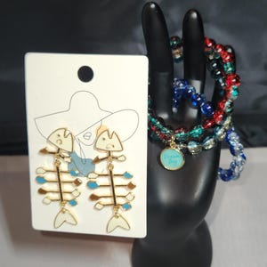 May include: A set of gold fishbone earrings with blue and white enamel accents. The earrings are displayed on a white card with a drawing of a woman wearing a hat. A set of three beaded bracelets with red, blue, and green beads are displayed on a black hand. The bracelets have a gold charm with the words "Dream Big" on it.