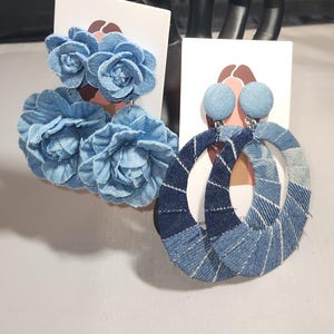 May include: Two pairs of denim earrings. One pair features large, light blue fabric flowers. The other pair has large, oval hoops made of denim fabric in shades of blue. Both pairs are displayed on white cards.